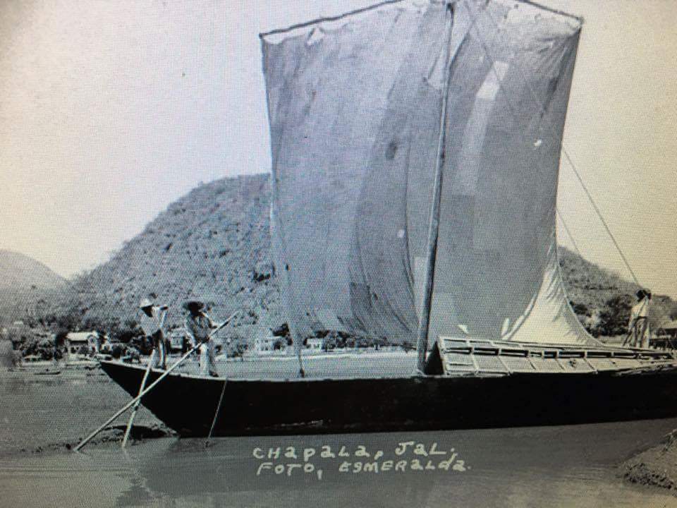 La imagen muestra un bote de vela estacionado cerca de la orilla de el lago de chapala, dado el paisaje circundante. Hay dos personas visibles a bordo del bote. Una de ellas está de pie en la proa del bote sosteniendo una vara o un remo, y la otra persona está un poco más atrás, parcialmente oscurecida por la vela. La vela es grande y cuadrada y parece estar un poco desgastada o arrugada, no está completamente desplegada. La escena es tranquila; el bote parece no estar en movimiento y el agua está calma. Hay una colina o montaña baja detrás del bote que domina el paisaje. La vegetación en la colina es dispersa, y parece ser un ambiente seco o semidesértico. En la parte frontal del bote, en la parte inferior de la imagen, hay un texto que parece ser un título o una referencia a la ubicación donde se tomó la foto: CHAPALA, JAL seguido de FOTO, ESMERALDA. Esto sugiere que la escena probablemente se desarrolle cerca de o en el Lago de Chapala, que está ubicado en el estado de Jalisco, México. El estilo de la foto y la calidad de la imagen sugieren que podría ser una foto antigua o histórica. El uso del blanco y negro contribuye a la sensación de que la foto podría ser de una época pasada, probablemente antes de la era del color en la fotografía de uso común.