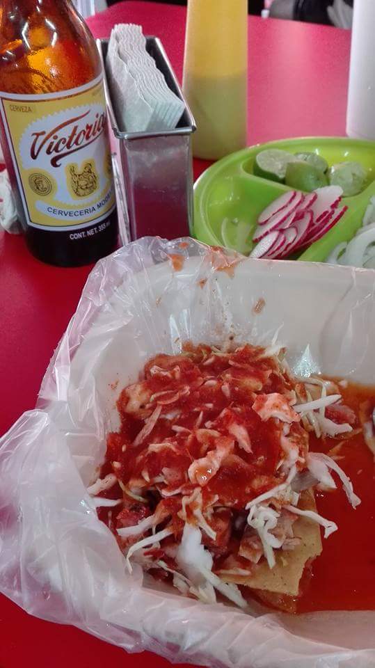 La imagen muestra una comida típica mexicana de la ciudad de Guadalajara. Al frente y en el centro, hay un plato que contiene lo que es una torta ahogada: un bollo de birote, un tipo de pan crujiente y ligeramente crujiente por fuera pero suave por dentro, relleno de carnitas de cerdo sazonadas con especias tradicionales cubiertos con salsa de tomate roja y espolvoreados con cebolla blanca picada. El plato luce desordenado y hay una bolsa de plástico utilizada como mantel o para mantener limpio el plato tradicional. A la izquierda de la comida se ve una botella de cerveza con la etiqueta Victoria que indica que es una marca de cerveza mexicana. Justo detrás de la botella de cerveza hay una servilletero de metal con servilletas blancas. Detrás del plato principal hay un recipiente pequeño de plástico de color verde con lo que parecen ser rodajas de limón y cebolla, lo que podría servir como aderezo o acompañamiento para la comida. Al fondo y hacia la derecha, puede apreciarse una botella de plástico amarillo con un líquido dentro que podría ser un condimento picante o salsa, muy común en la gastronomía mexicana, y también hay un vaso blanco que, aunque no está enfocado, podría contener alguna bebida.